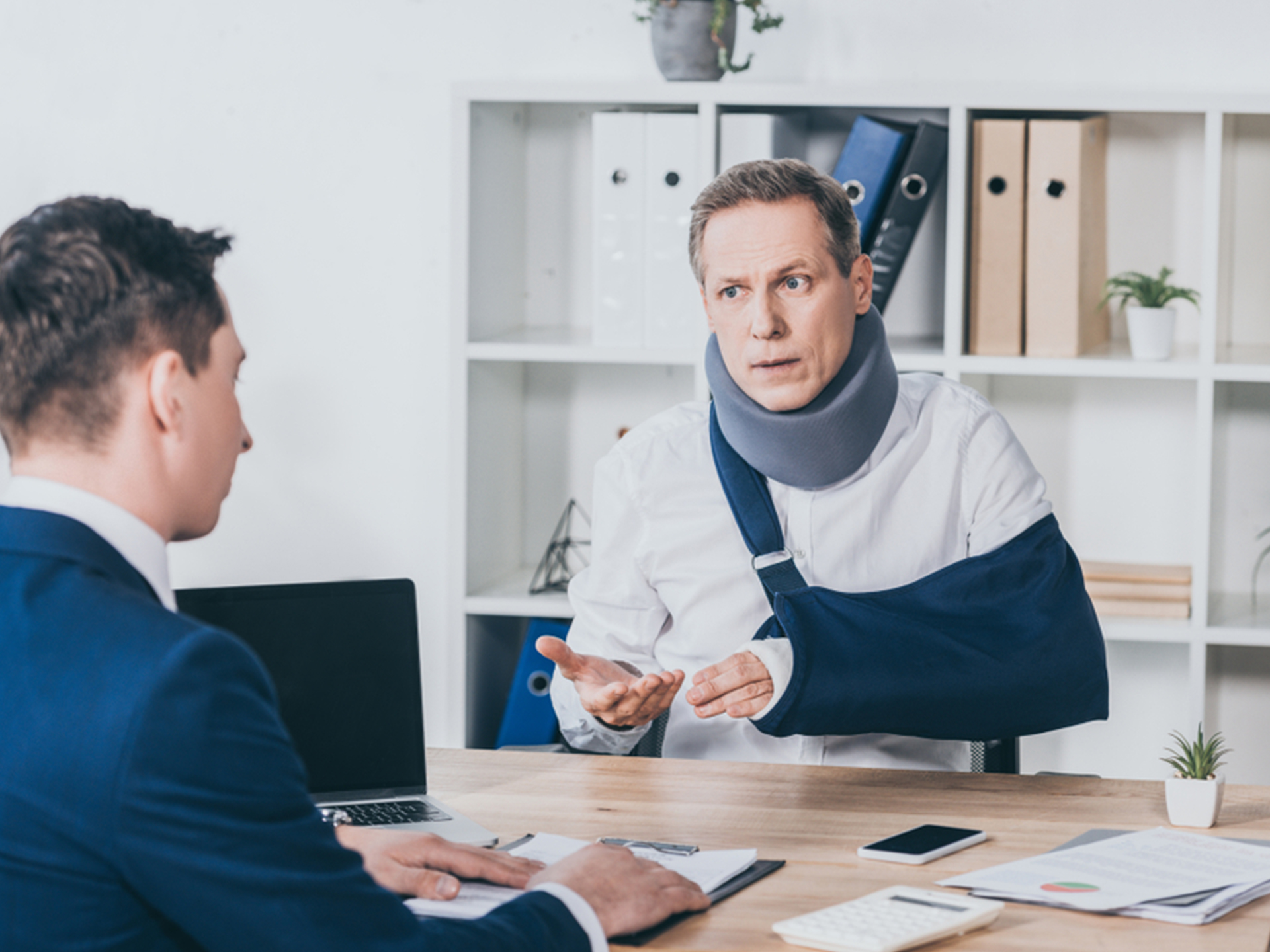 middle aged worker in neck brace with broken arm sitting at table and talking to businessman in blue jacket in office