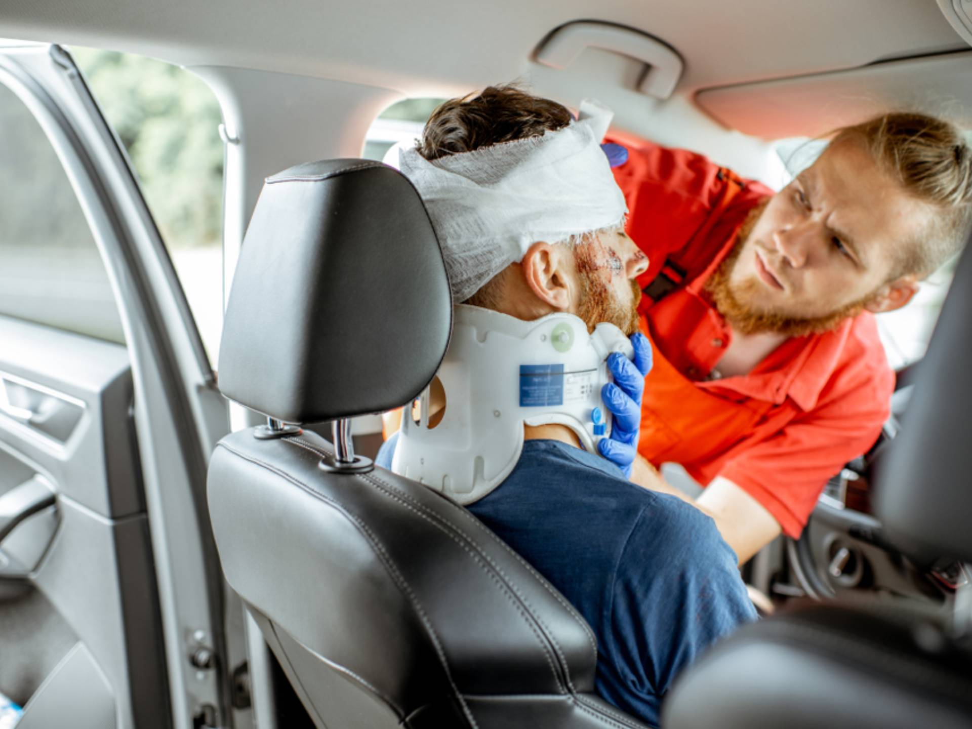 Medic applying bandage on the head of victim with serious damages sitting on the driver seat after the road accident
